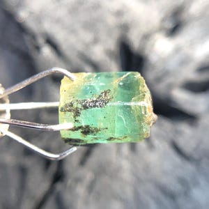 May include: A green emerald gemstone with a rough, natural finish. The emerald is held in a metal clamp.