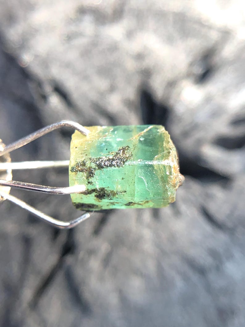 May include: A green emerald gemstone with a rough, natural finish. The emerald is held in a metal clamp.