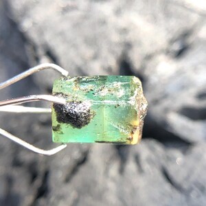 May include: A green emerald gemstone with black inclusions, held by tweezers. The emerald is cut into a rectangular shape with sharp edges.
