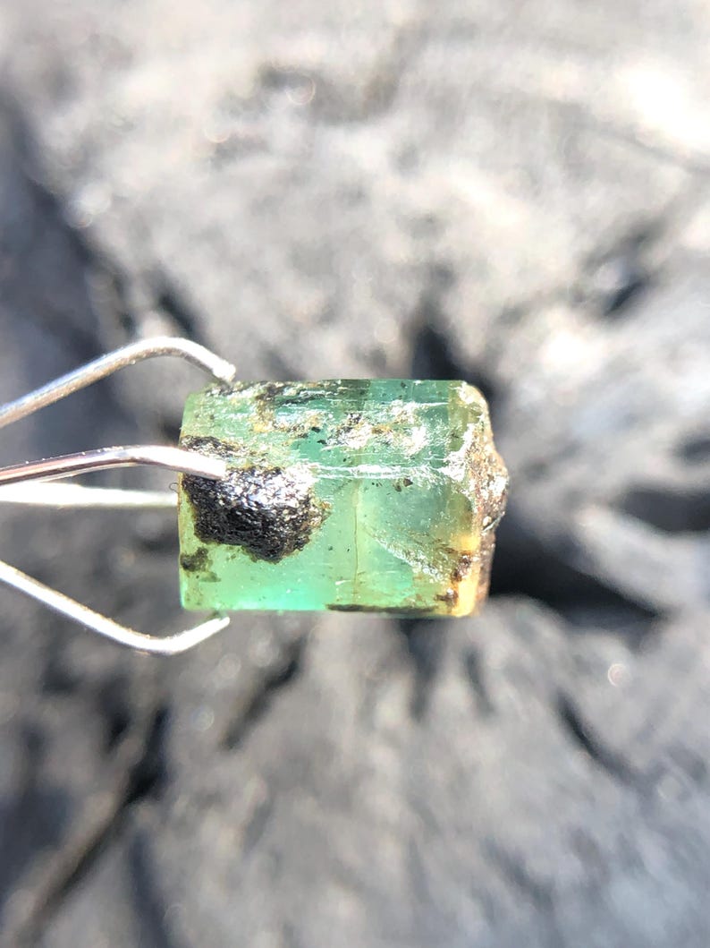May include: A green emerald gemstone with black inclusions, held by tweezers. The emerald is cut into a rectangular shape with sharp edges.