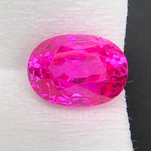Può includere: Una gemma dal taglio ovale vibrante con un brillante colore rosa acceso. La pietra presenta numerose superfici sfaccettate che riflettono la luce, creando un aspetto scintillante. La gemma è posizionata su una superficie testurizzata color bianco sporco.