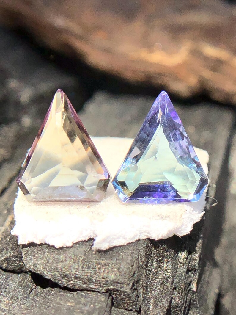 May include: Two triangular faceted gemstones, one is a pale yellow-green with a hint of purple, the other is a light blue with a hint of green.