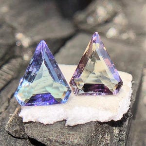 May include: Two triangular faceted gemstones, one is a light blue with a purple hue, the other is a light yellow with a purple hue.