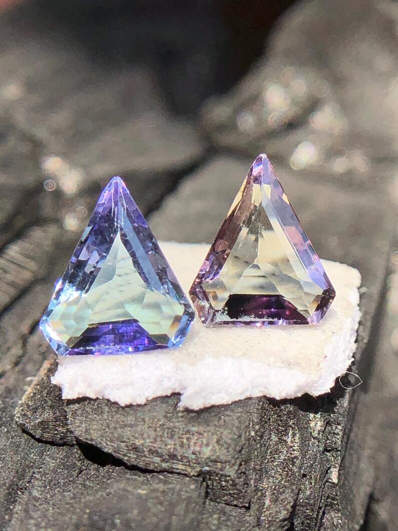 May include: Two triangular faceted gemstones, one is a light blue with a purple hue, the other is a light yellow with a purple hue.