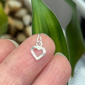 May include: A silver charm in the shape of a heart with a textured design. The heart is hollow and has a small loop at the top for attaching to a chain or bracelet.