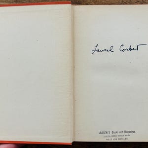 May include: Open vintage book with orange cover and off-white pages. The right page features a handwritten signature, "Laurel Corbett." The bottom of the page has the text "LARSEN'S - Books and Magazines" and address details.