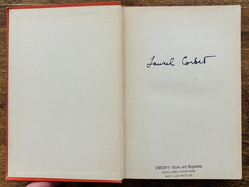 May include: Open vintage book with orange cover and off-white pages. The right page features a handwritten signature, "Laurel Corbett." The bottom of the page has the text "LARSEN'S - Books and Magazines" and address details.