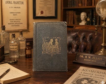 May include: A vintage book with a dark blue cover and gold embossed illustration of two figures. The book is on a wooden desk with other antique items, including glass bottles, a desk nameplate, and a document titled "Mesmerism".
