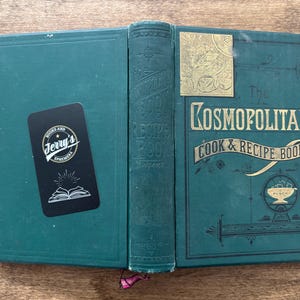May include: An open, antique cookbook titled "The Kosmopolitan Cook & Recipe Book" in teal green with gold lettering. A black sticker with white text and a book illustration is on the left cover. The book is on a wooden surface.