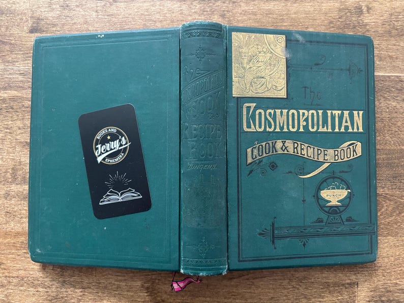 May include: An open, antique cookbook titled "The Kosmopolitan Cook & Recipe Book" in teal green with gold lettering. A black sticker with white text and a book illustration is on the left cover. The book is on a wooden surface.