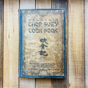 Pu&ograve; includere: Un libro di cucina antico intitolato "Mandarin Chop Suey Cook Book" con una copertina dorata e blu sbiadita. La copertina presenta testo ornato e caratteri cinesi. Il titolo e la descrizione del libro sono visibili, con il prezzo indicato come "One Dollar".