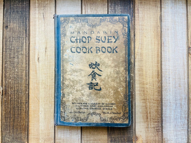 Pu&ograve; includere: Un libro di cucina antico intitolato "Mandarin Chop Suey Cook Book" con una copertina dorata e blu sbiadita. La copertina presenta testo ornato e caratteri cinesi. Il titolo e la descrizione del libro sono visibili, con il prezzo indicato come "One Dollar".