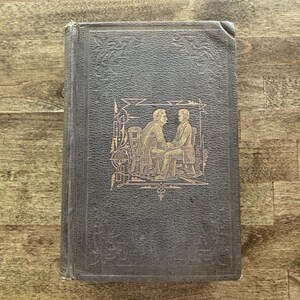 Puede incluir: Un libro antiguo con una cubierta gris oscuro y patrones florales en relieve. La portada presenta una ilustraci&oacute;n dorada de dos hombres sentados en sillas. Los bordes del libro muestran signos de desgaste, lo que sugiere su antig&uuml;edad.