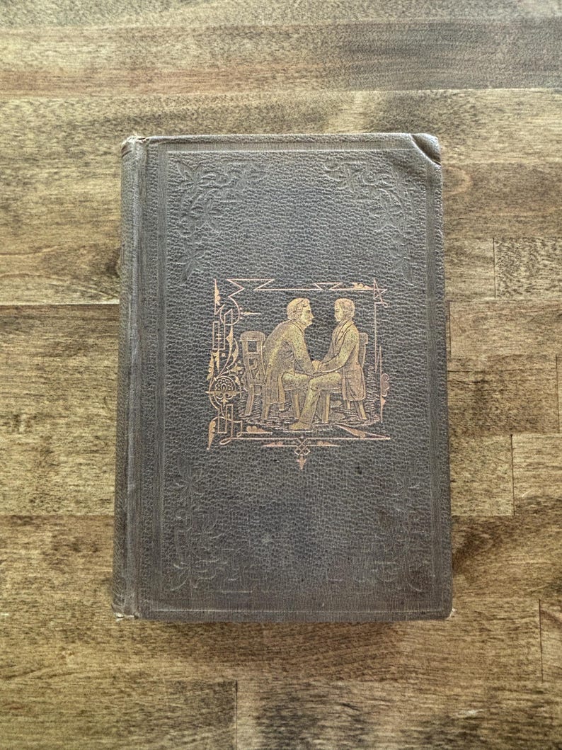 Puede incluir: Un libro antiguo con una cubierta gris oscuro y patrones florales en relieve. La portada presenta una ilustraci&oacute;n dorada de dos hombres sentados en sillas. Los bordes del libro muestran signos de desgaste, lo que sugiere su antig&uuml;edad.