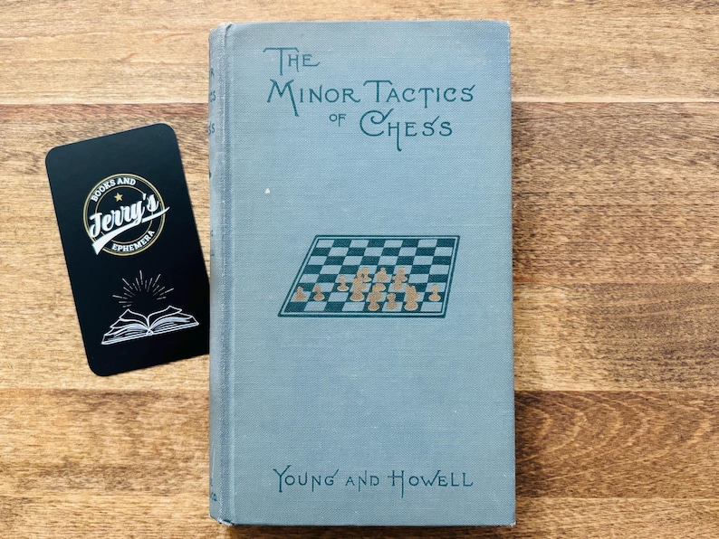 May include: A vintage book titled "The Minor Tactics of Chess" with a light blue cover. The cover features a chess board illustration and the authors' names, Young and Howell. A black business card with the text "Books and Jerry's Ephemera" is next to the book.