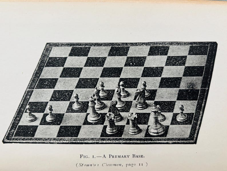 May include: An illustration of a chessboard with chess pieces in a starting position. The board is black and white checkered, with a dark border. The chess pieces are detailed and arranged for the start of a game. Text at the bottom reads "FIG. 1. - A PRIMARY BASE."