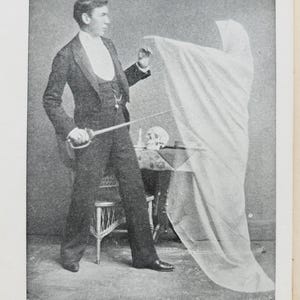 May include: A vintage spirit photograph in black and white depicts a man in a suit holding a sword, with a draped figure and a skull on a table. The text at the bottom reads "SPIRIT PHOTOGRAPH. [Taken by the Author.]"