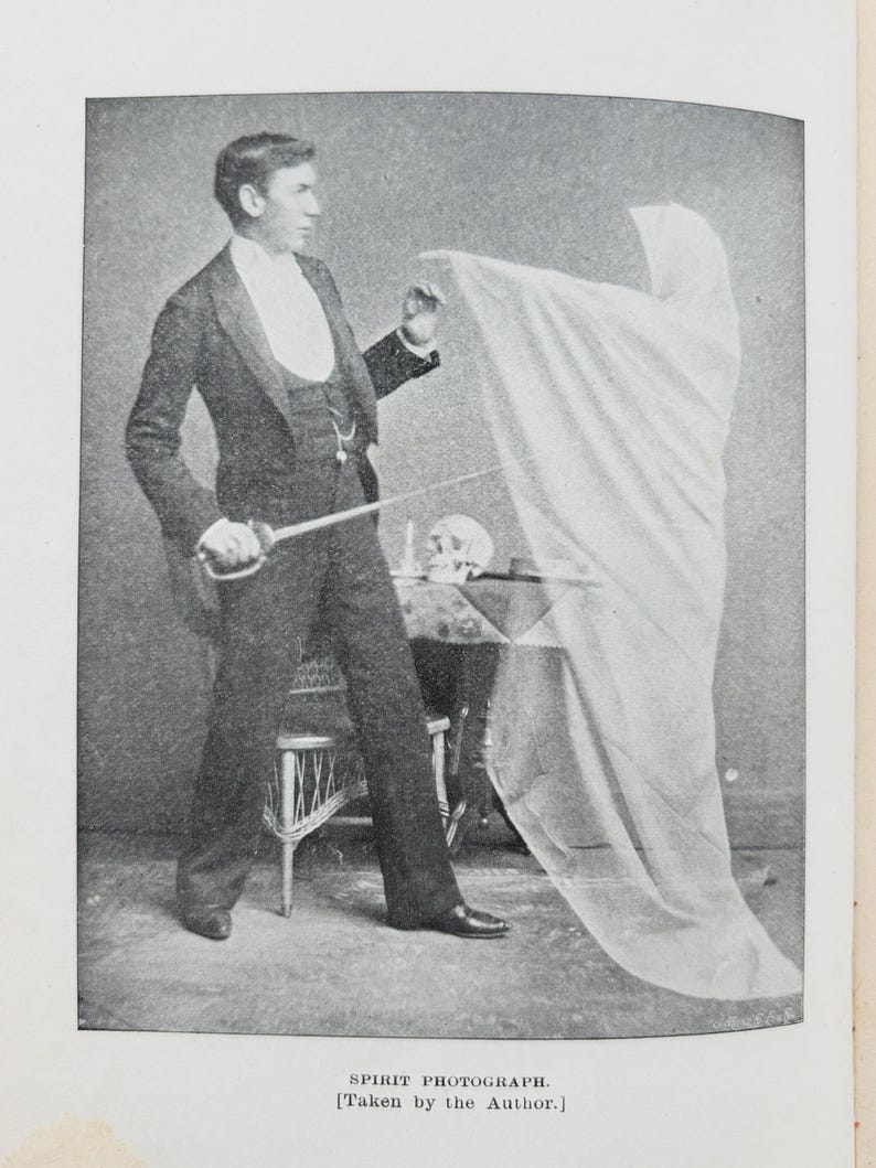 May include: A vintage spirit photograph in black and white depicts a man in a suit holding a sword, with a draped figure and a skull on a table. The text at the bottom reads "SPIRIT PHOTOGRAPH. [Taken by the Author.]"