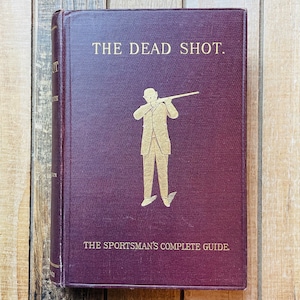 May include: A burgundy book with gold lettering titled "The Dead Shot. The Sportsman's Complete Guide." The cover features a silhouette of a person holding a rifle.