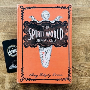 May include: An orange book titled "The Spirit World Unmasked" with a ghost illustration on the cover. A black card with the text "Books and Jerry's Ephemera" is next to the book. The book is on a wooden surface.