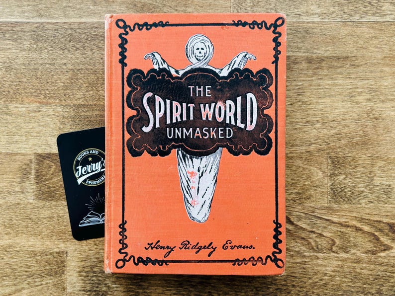 May include: An orange book titled "The Spirit World Unmasked" with a ghost illustration on the cover. A black card with the text "Books and Jerry's Ephemera" is next to the book. The book is on a wooden surface.