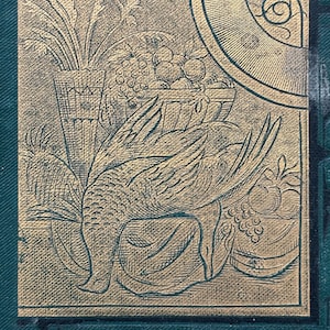 May include: A detailed gold-toned illustration on a dark green background. The image features a bird with outstretched wings, a basket of fruit, and a vase of plants. The design has a vintage, engraved appearance.