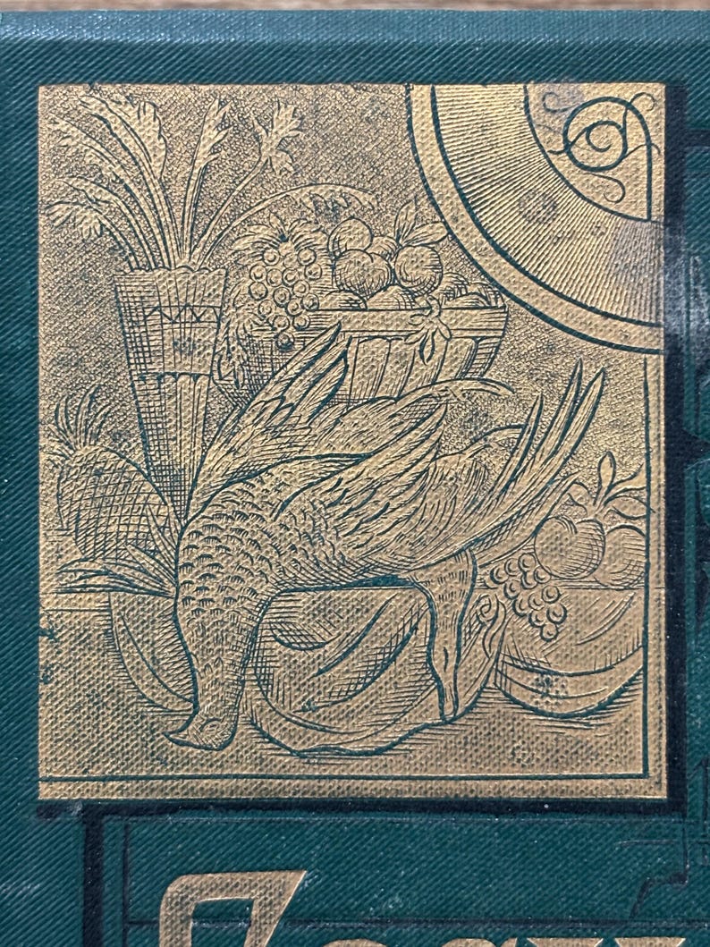 May include: A detailed gold-toned illustration on a dark green background. The image features a bird with outstretched wings, a basket of fruit, and a vase of plants. The design has a vintage, engraved appearance.