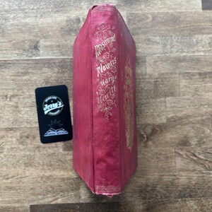 May include: A vintage book with a red cover and gold lettering. The spine features the title "Mrs. Hemans' Flowers" and the author's name, Mary Howitt. A black card with the text "Jerry's" is next to the book.