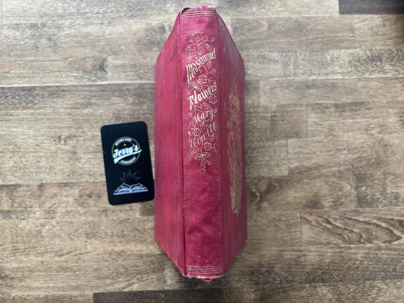 May include: A vintage book with a red cover and gold lettering. The spine features the title "Mrs. Hemans' Flowers" and the author's name, Mary Howitt. A black card with the text "Jerry's" is next to the book.