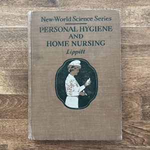 May include: A vintage book titled "Personal Hygiene and Home Nursing" from the New-World Science Series. The cover is tan with green text and an illustration of a woman in a nurse's uniform. The book is on a wooden surface.