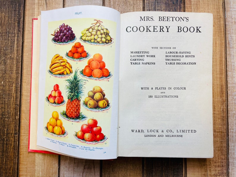 Pode incluir: Um livro de cozinha vintage aberto intitulado "Mrs. Beeton's Cookery Book" com se&ccedil;&otilde;es sobre marketing, trabalho de lavanderia, talha, guardanapos de mesa, economia de trabalho, dicas para o lar, amarra&ccedil;&atilde;o e decora&ccedil;&atilde;o de mesa. O livro apresenta 8 placas em cores e 150 ilustra&ccedil;&otilde;es. A capa mostra uma ilustra&ccedil;&atilde;o colorida de v&aacute;rias frutas, incluindo uvas, bananas, laranjas, ma&ccedil;&atilde;s e um abacaxi.