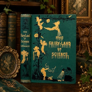 May include: A hardcover book titled The Fairy-Land of Science by A.B. Buckley with a dark green cover featuring gold embossed illustrations of winged figures, vines, and a figure holding a lantern, with matching gold text on the book spine.