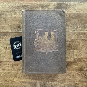 Puede incluir: Un libro antiguo con una cubierta gris oscuro e ilustraciones doradas en relieve de dos figuras. Una tarjeta de visita negra con el texto "Jerry's Books and Ephemera" est&aacute; al lado del libro.