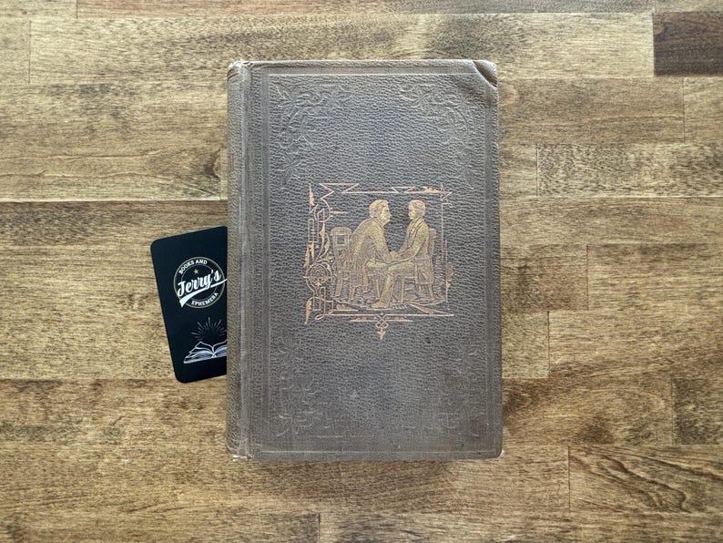 Puede incluir: Un libro antiguo con una cubierta gris oscuro e ilustraciones doradas en relieve de dos figuras. Una tarjeta de visita negra con el texto "Jerry's Books and Ephemera" est&aacute; al lado del libro.