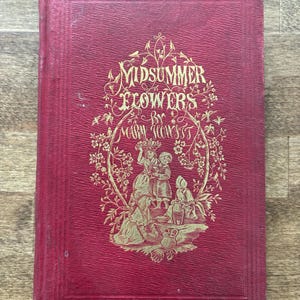 May include: A vintage book titled "Midsummer Flowers" by Mary Howitt. The cover is a deep red with gold embossed lettering and illustrations of figures and floral elements. The book has a textured appearance.