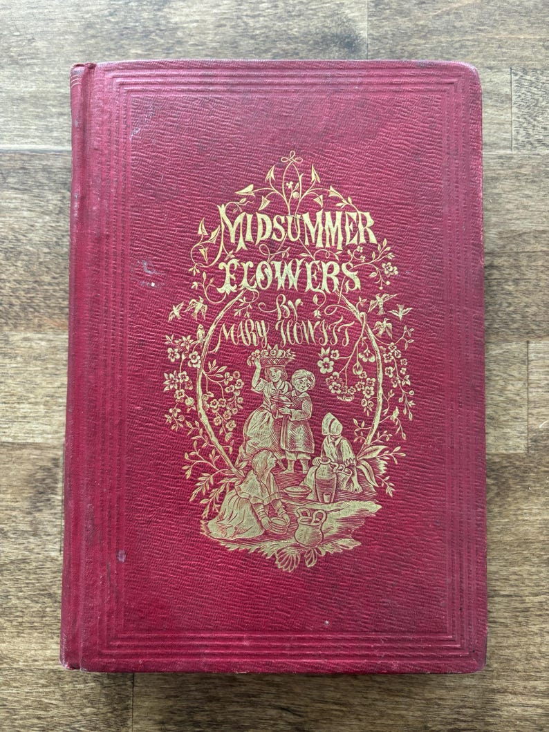 May include: A vintage book titled "Midsummer Flowers" by Mary Howitt. The cover is a deep red with gold embossed lettering and illustrations of figures and floral elements. The book has a textured appearance.