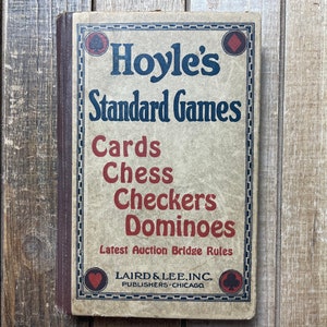 Puede incluir: Un libro vintage titulado "Hoyle's Standard Games" con una cubierta marr&oacute;n y texto rojo y azul. El libro presenta una lista de juegos, incluyendo cartas, ajedrez, damas y domin&oacute;. El libro tambi&eacute;n incluye el texto "Latest Auction Bridge Rules" y la informaci&oacute;n del editor "LAIRD & LEE, INC. PUBLISHERS-CHICAGO".