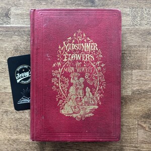 May include: A vintage book titled "Midsummer Flowers" by Mary Howitt, with a red cover and gold lettering and illustrations. A black card with white text is next to the book. The book is on a wooden surface.