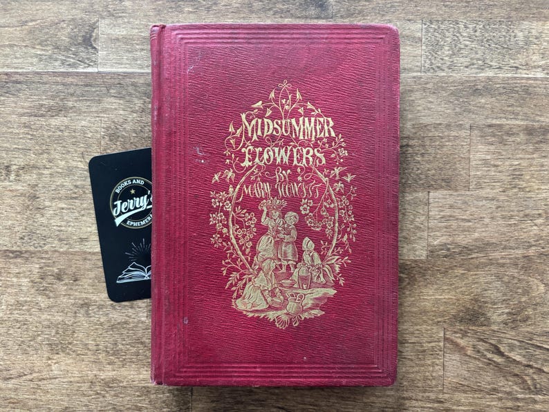 May include: A vintage book titled "Midsummer Flowers" by Mary Howitt, with a red cover and gold lettering and illustrations. A black card with white text is next to the book. The book is on a wooden surface.