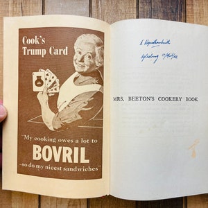Pode incluir: Um livro de cozinha vintage intitulado "Mrs. Beeton's Cookery Book" com um an&uacute;ncio em marrom e branco para Bovril na capa. O an&uacute;ncio apresenta uma mulher segurando um baralho de cartas e o texto "Cook's Trump Card" e "My cooking owes a lot to Bovril - so do my nicest sandwiches."
