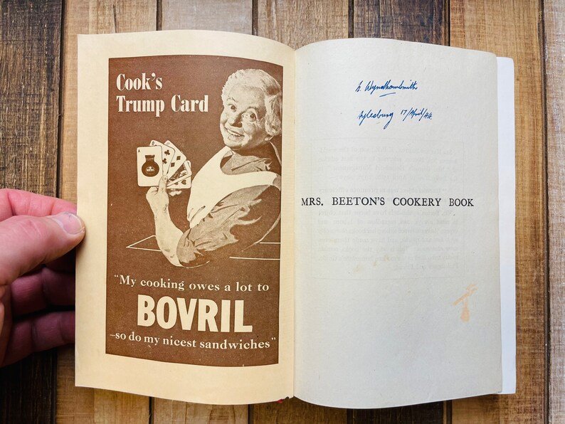 Pode incluir: Um livro de cozinha vintage intitulado "Mrs. Beeton's Cookery Book" com um an&uacute;ncio em marrom e branco para Bovril na capa. O an&uacute;ncio apresenta uma mulher segurando um baralho de cartas e o texto "Cook's Trump Card" e "My cooking owes a lot to Bovril - so do my nicest sandwiches."