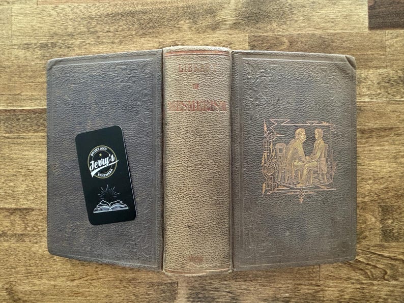 Puede incluir: Un libro antiguo abierto con una cubierta desgastada y texturizada, con el t&iacute;tulo "Library of Esoteric". Una tarjeta de visita negra con el texto "Jerry's Ephemera" descansa sobre el libro. La cubierta del libro es de color gris oscuro.