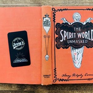 May include: An open vintage book with an orange cover and spine. The title "The Spirit World Unmasked" is displayed on the cover in a decorative font, with a ghostly illustration. A black business card is on the left page.