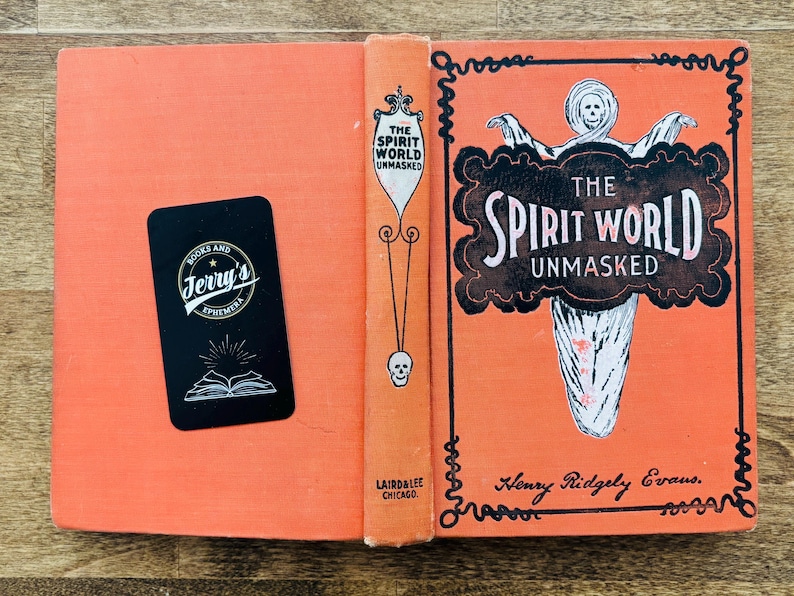 May include: An open vintage book with an orange cover and spine. The title "The Spirit World Unmasked" is displayed on the cover in a decorative font, with a ghostly illustration. A black business card is on the left page.