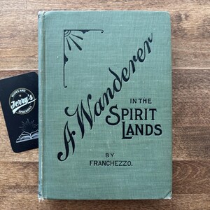 May include: A vintage book titled "A Wanderer in the Spirit Lands" by Franchezzo, in a muted green cover. A black business card from Jerry's Books and Ephemera is next to the book on a wooden surface.