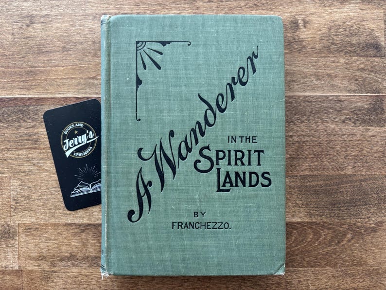 May include: A vintage book titled "A Wanderer in the Spirit Lands" by Franchezzo, in a muted green cover. A black business card from Jerry's Books and Ephemera is next to the book on a wooden surface.