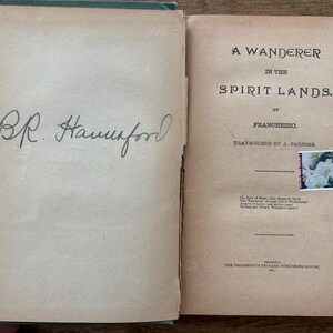 May include: Open book with aged, tan pages. The left page has a handwritten signature in black ink. The right page displays the title "A Wanderer in the Spirit Lands" by Franchezzo. A small stamp with white flowers is affixed.
