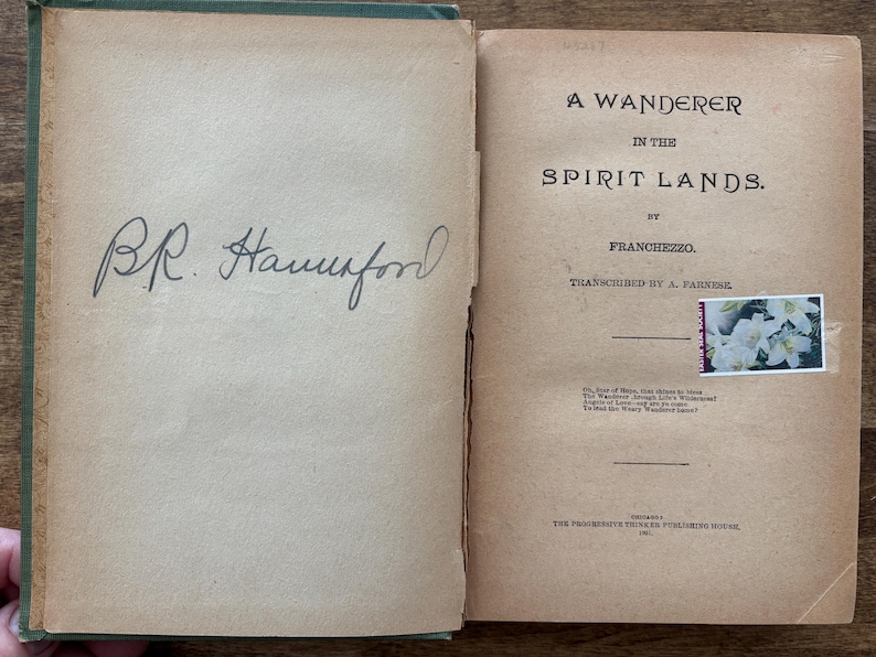 May include: Open book with aged, tan pages. The left page has a handwritten signature in black ink. The right page displays the title "A Wanderer in the Spirit Lands" by Franchezzo. A small stamp with white flowers is affixed.