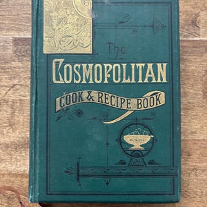 May include: Antique cookbook titled "The Cosmopolitan Cook & Recipe Book" with a dark teal cover and gold lettering. The cover features decorative gold accents and a bookmark. The book is on a wooden surface.