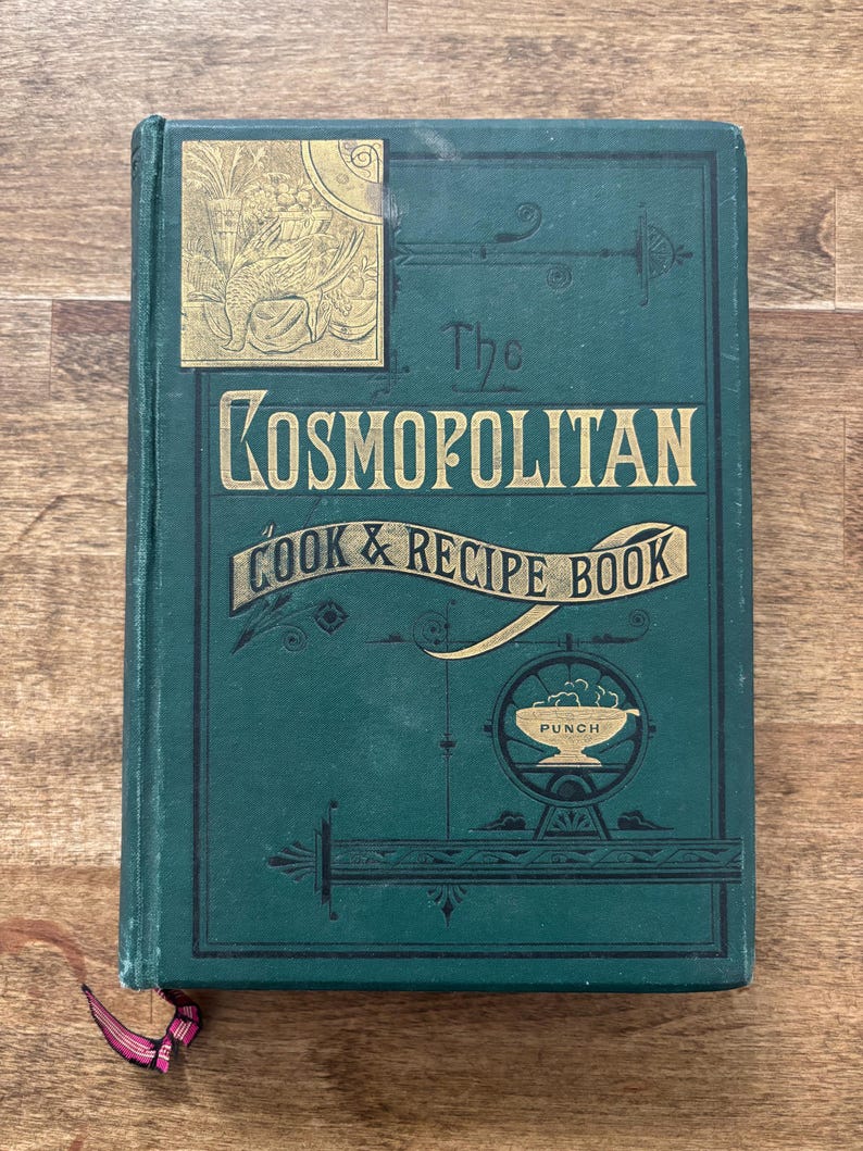 May include: Antique cookbook titled "The Cosmopolitan Cook & Recipe Book" with a dark teal cover and gold lettering. The cover features decorative gold accents and a bookmark. The book is on a wooden surface.